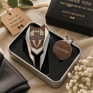 Könnte beinhalten: Ein Golf-Pitchgabel aus silberfarbenem Metall und dunklem Holz sowie ein runder magnetischer Ballmarker in einer Metalldose. Die Pitchgabel zeigt ein Smoking-Motiv mit dem Text BESTMAN, der Ballmarker ist mit dem Namen RICHARD graviert.