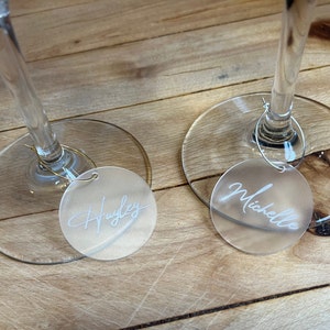 Puede incluir: Dos marcadores de copa circulares de acr&iacute;lico esmerilado unidos a finos aros de metal. Cada marcador presenta un nombre grabado en letra cursiva blanca, uno dice Hayley y el otro Michelle, colocados en la base de dos copas de vino.