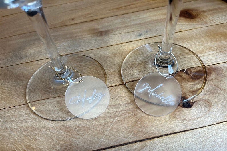 Puede incluir: Dos marcadores de copa circulares de acr&iacute;lico esmerilado unidos a finos aros de metal. Cada marcador presenta un nombre grabado en letra cursiva blanca, uno dice Hayley y el otro Michelle, colocados en la base de dos copas de vino.