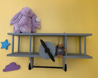 May include: A gray wooden airplane shelf with a purple bunny and a gray teddy bear on it. The shelf has a black propeller and a black metal rod for hanging items.