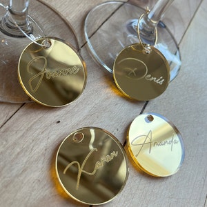 Puede incluir: Cuatro dijes circulares para copas de vino hechos de acr&iacute;lico con espejo dorado y nombres grabados. Dos dijes est&aacute;n unidos a las bases de vidrio transparente, mientras que dos est&aacute;n sueltos. Los nombres Joanne, Denis, Karan y Amanda est&aacute;n grabados.