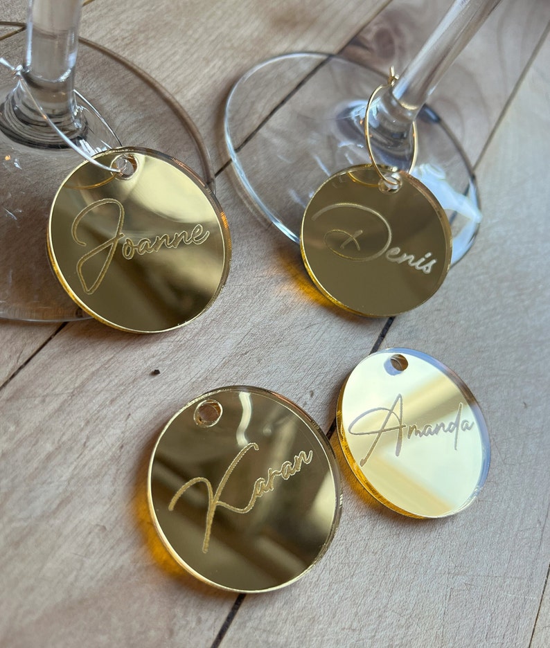 Puede incluir: Cuatro dijes circulares para copas de vino hechos de acr&iacute;lico con espejo dorado y nombres grabados. Dos dijes est&aacute;n unidos a las bases de vidrio transparente, mientras que dos est&aacute;n sueltos. Los nombres Joanne, Denis, Karan y Amanda est&aacute;n grabados.