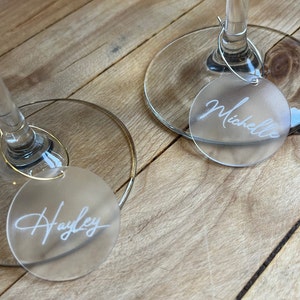 Puede incluir: Dos marcadores de copa circulares de acr&iacute;lico esmerilado unidos a los tallos de bases de vidrio transparente con aros de metal finos. Cada marcador presenta un nombre grabado en letra cursiva blanca, espec&iacute;ficamente Hayley y Michelle.