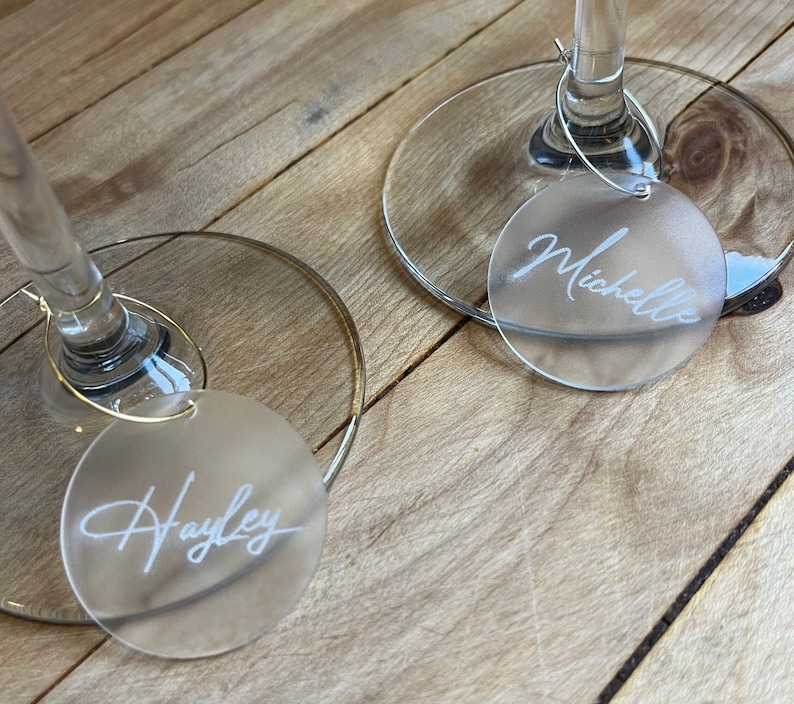 Puede incluir: Dos marcadores de copa circulares de acr&iacute;lico esmerilado unidos a los tallos de bases de vidrio transparente con aros de metal finos. Cada marcador presenta un nombre grabado en letra cursiva blanca, espec&iacute;ficamente Hayley y Michelle.