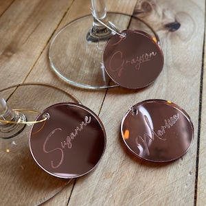 Puede incluir: Tres dijes circulares para copas de vino hechos de acr&iacute;lico con acabado de espejo en oro rosa, grabados con los nombres Grayson, Suzanne y Marissa en letra cursiva. Cada dije incluye un fino aro de alambre dorado para el tallo.
