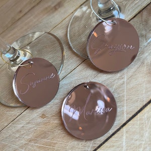 Puede incluir: Tres dijes circulares para copas de vino hechos de acr&iacute;lico con acabado de espejo en oro rosa, cada uno con un nombre grabado en letra cursiva. Dos dijes est&aacute;n sujetos a las bases de copas de cristal, mientras que uno est&aacute; separado. Los nombres son Suzanne, Grayson y Marissa.