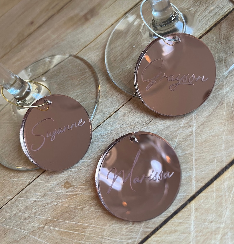 Puede incluir: Tres dijes circulares para copas de vino hechos de acr&iacute;lico con acabado de espejo en oro rosa, cada uno con un nombre grabado en letra cursiva. Dos dijes est&aacute;n sujetos a las bases de copas de cristal, mientras que uno est&aacute; separado. Los nombres son Suzanne, Grayson y Marissa.