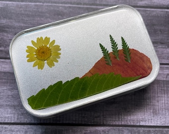 May include: A rectangular silver metal tin featuring a decorative landscape design on the lid. The design is composed of pressed botanical elements, including a yellow flower, a large green leaf, a reddish-brown leaf, and three small fern fronds.