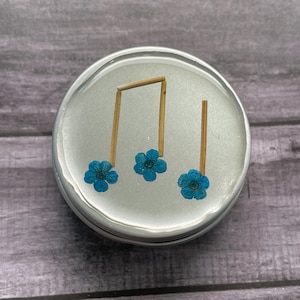 May include: A round, silver-colored container with a clear, resin-like top. Inside, a musical note is crafted from small wooden pieces and three small, vibrant blue flowers. The container sits on a gray wooden surface.