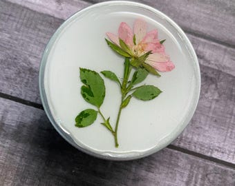 May include: A circular resin coaster featuring a pressed pink rose and green leaves. The flower is set on a white base, encased in clear resin, and has a silver edge. A decorative floral item.