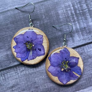 May include: A pair of round wooden earrings featuring pressed purple delphinium flowers on the front surface. Each earring is attached to a silver-toned metal fishhook ear wire, showcasing the natural wood grain and the vibrant petals of the flower.