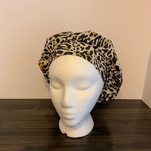 May include: A satin bonnet with a leopard print design in black, tan, and cream colors. The bonnet is displayed on a white foam mannequin head, set against a neutral background.