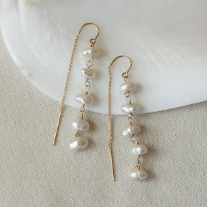 Puede incluir: Un par de pendientes colgantes dorados. Cada pendiente presenta una cadena con una serie de perlas blancas de forma irregular. Los pendientes se muestran sobre una superficie blanca texturizada, ideal para joyería o accesorios.