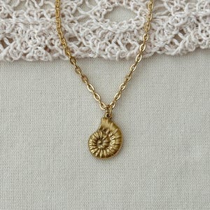 May include: A gold-colored necklace featuring a pendant shaped like a nautilus shell. The pendant is suspended from a delicate chain. The necklace is displayed on a cream-colored, textured surface.