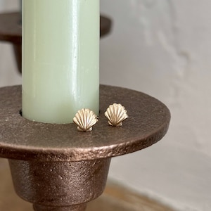 May include: A pair of small gold stud earrings shaped like seashells with detailed ridges. The earrings are resting on the flat, circular, textured bronze-colored metal base of a candle holder that holds a pale green taper candle.
