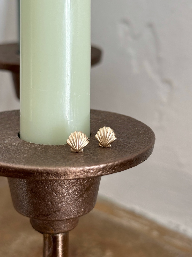 May include: A pair of small gold stud earrings shaped like seashells with detailed ridges. The earrings are resting on the flat, circular, textured bronze-colored metal base of a candle holder that holds a pale green taper candle.
