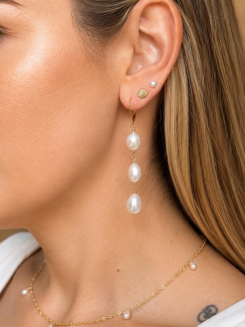 May include: A gold drop earring featuring three vertically aligned white teardrop-shaped pearls suspended from a gold wire hook. The earring is worn alongside a small gold shell-shaped stud and a tiny white pearl stud earring.