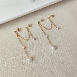 May include: A pair of gold-tone chain earrings featuring a stud post with a small ball accent. Each earring has two delicate gold chains of varying lengths, each finished with a small white pearl bead at the end and one additional pearl.