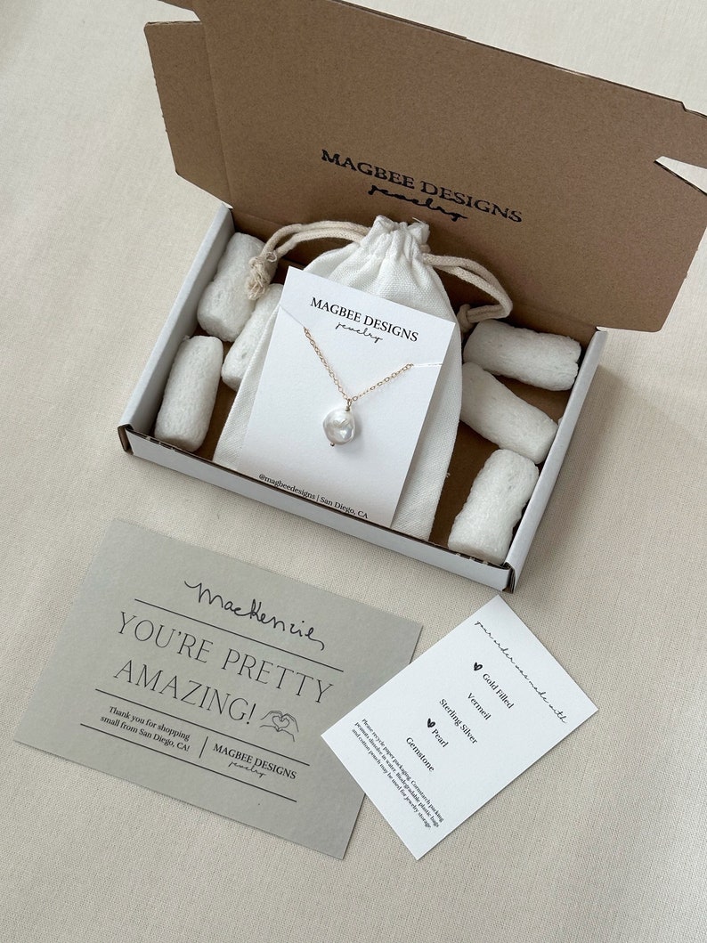 May include: A gold chain necklace with a single white baroque pearl pendant on a display card inside a cardboard box with white packing peanuts and a drawstring pouch. Includes a thank you note and a card listing materials like gold filled.
