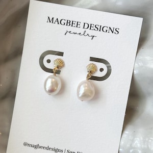 May include: A pair of pearl earrings on a white card with the text "MAGBEE DESIGNS jewelry". The earrings have a gold seashell design at the top and a large, white, oval pearl. The card also includes the text "@magbeedesigns | San Diego, CA".