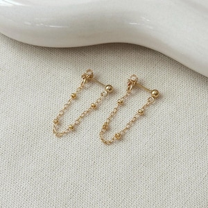 May include: A pair of gold-toned stud earrings featuring a delicate chain that connects the post to the back. Each earring has a small ball stud and a fine link chain with evenly spaced tiny gold beads, creating a looped drop design.