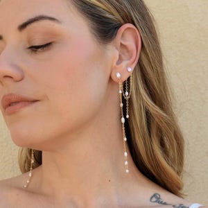 May include: A gold chain earring featuring multiple small white freshwater pearls spaced along the length of the chain. The earring connects two points on the earlobe with a draped chain design, creating a long, dangling effect with pearls.