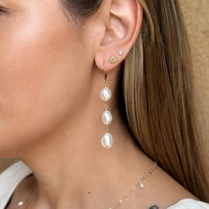 May include: A gold drop earring featuring three white baroque pearls suspended in a vertical line. The ear also displays a small gold seashell stud and a tiny white pearl stud. A delicate gold chain necklace with small white pearls is visible.