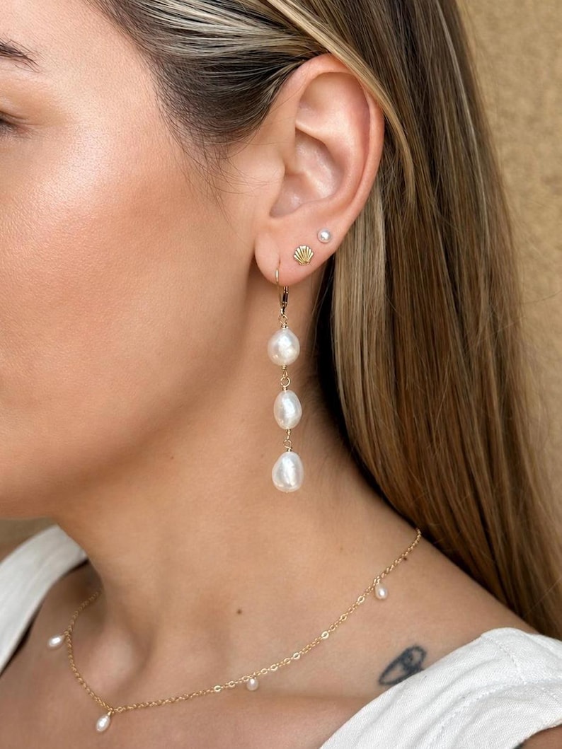 May include: A gold drop earring featuring three white baroque pearls suspended in a vertical line. The ear also displays a small gold seashell stud and a tiny white pearl stud. A delicate gold chain necklace with small white pearls is visible.