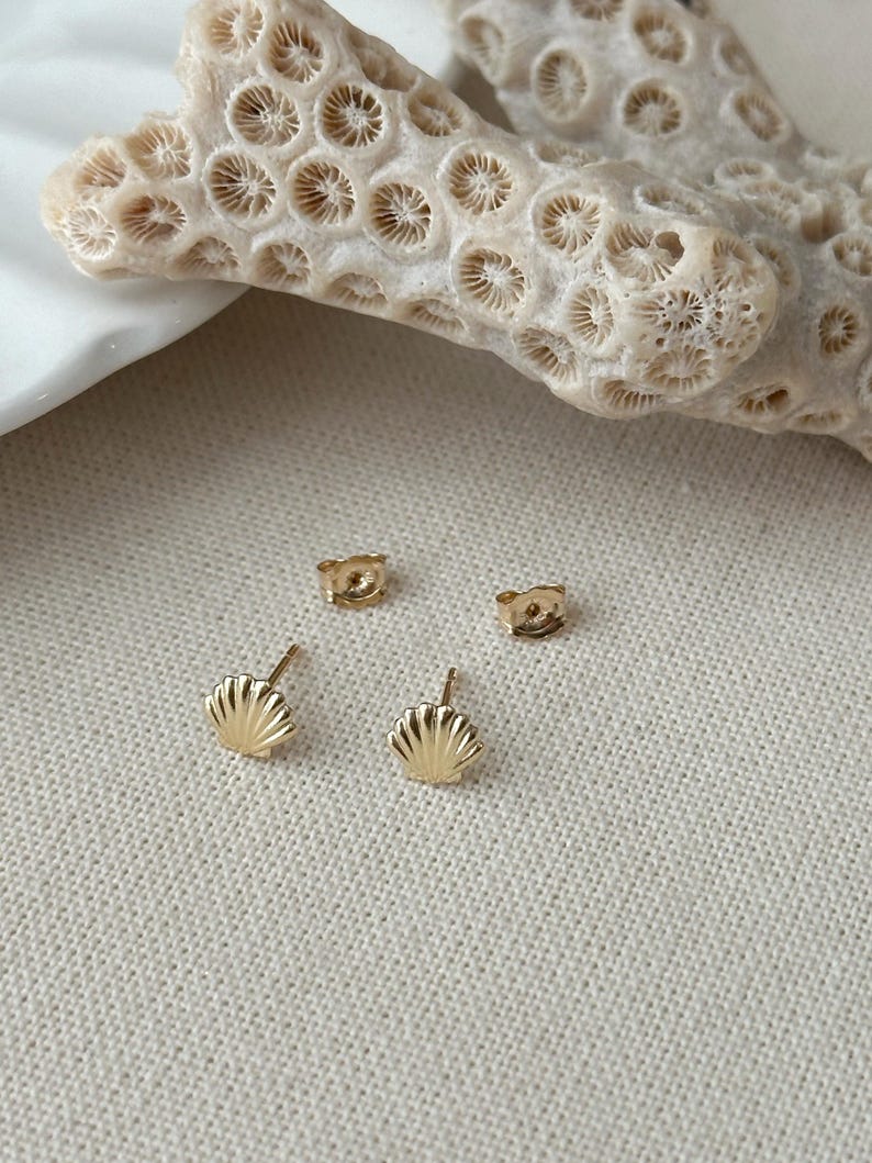 May include: A pair of gold stud earrings shaped like small seashells, each featuring a textured fan design with vertical ridges. The earrings are accompanied by two matching gold butterfly-style earring backs, positioned separately from the studs.