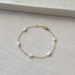 Könnte beinhalten: Ein zartes Armband aus goldfarbener Kette mit sechs ovalen, weißen Perlen. Das Armband hat einen kleinen Verschluss und wird auf einer neutralfarbenen Oberfläche präsentiert. Der Schmuck ist schlicht und elegant.