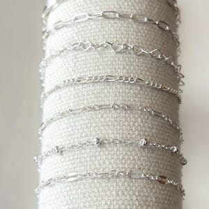 Könnte beinhalten: Eine Sammlung silberner Kettenarmbänder, präsentiert auf einem neutralen Stoffhintergrund. Die Armbänder weisen verschiedene Gliederarten auf, darunter ovale, herzförmige und perlenbesetzte Designs. Die Ketten sind zart und glänzend und reflektieren das Licht.