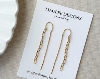 May include: A pair of gold-toned threader earrings featuring a delicate chain design with elongated rectangular links. The earrings are displayed on a white rectangular card printed with the text MAGBEE DESIGNS jewelry and @magbeedesigns | San Diego, CA.