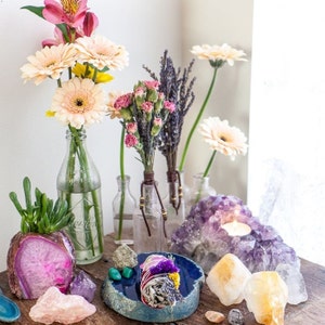 Puede incluir: Un arreglo de mesa con varios cristales, flores y un palo de limpieza. Los cristales incluyen amatista, cuarzo transparente y citrino. Las flores son margaritas blancas y rosadas, lavanda y claveles rosados. El palo de limpieza está envuelto en tela blanca y tiene flores de lavanda secas en la parte superior.