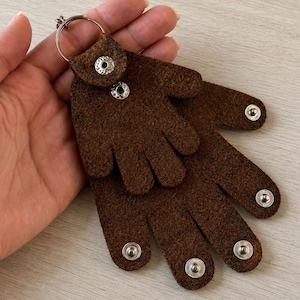 May include: A dark brown felt keychain shaped like two overlapping hands. The smaller hand is attached to a silver metal keyring and secured with a silver snap button. The larger hand features four additional silver snap buttons at the fingertips.