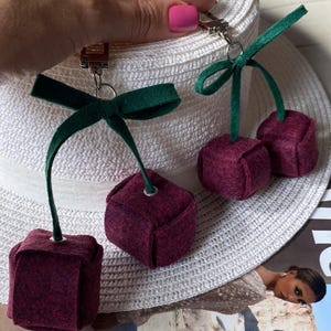 May include: Two cherry-shaped keychains made of dark maroon felt fabric. Each keychain features a pair of cube-shaped felt cherries attached to a green felt stem tied in a bow, connected to a silver-toned metal lobster clasp hardware.
