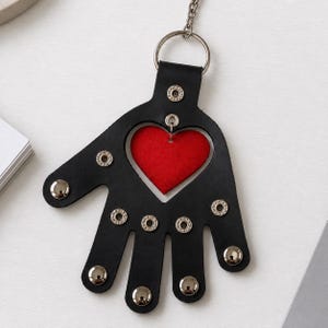 May include: A black hand-shaped keychain featuring a central red heart cutout. The black material is accented with silver-toned metal rivets and studs. A silver metal ring and chain are attached to the top of the hand-shaped piece.