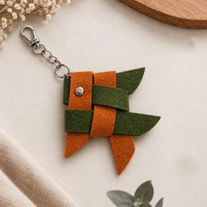 May include: A felt keychain featuring interwoven strips of orange and dark green fabric. The design is secured with a single silver-toned metal rivet and attached to a silver-colored metal lobster clasp and split ring for easy attachment.