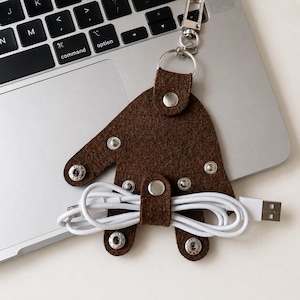 May include: A dark brown felt cable organizer shaped like a hand with silver metal snap buttons. The organizer features a silver metal swivel clasp at the top and holds a white USB charging cable wrapped horizontally across the center.
