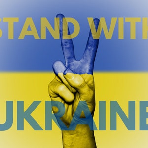 Op de afbeelding: Een hand die een vredesteken maakt, geschilderd in de kleuren van de Oekraïense vlag, met de woorden "STAND WITH UKRAINE" eroverheen. De achtergrond is een verloop van blauw en geel, dat de vlag weerspiegelt.