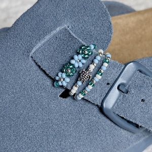 Puede incluir: Dos abalorios de cuentas sujetos a la tira de un zapato de ante azul. Uno presenta una hilera de pequeñas cuentas en forma de flor azul claro y verde oscuro. El otro consiste en una tira de cuentas azul claro, blancas y verdes con una tortuga plateada.