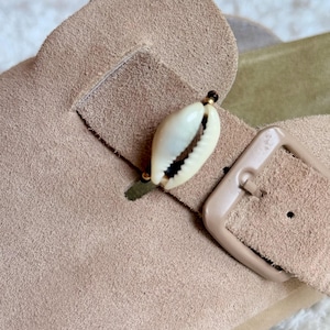 May include: A beige suede sandal featuring a single cowrie shell charm attached to the strap. The sandal includes a matching beige buckle and a light green footbed, showcasing a textured suede finish on the upper strap and a smooth buckle.