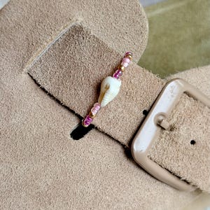 May include: A decorative charm featuring a small white seashell centered on a beaded strand of pink, purple, and metallic gold beads. The charm is attached to the strap of a tan suede sandal, which includes a matching tan buckle and hardware.