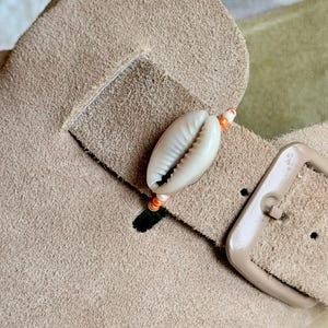 May include: A beige suede sandal strap featuring a single white cowrie shell centred on a thin string of small orange and white beads. The strap includes a matching beige buckle with a rectangular frame and a single prong for adjustment.