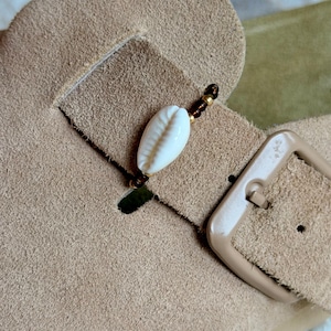 May include: A beige suede sandal strap featuring a decorative accessory made of a white cowrie shell flanked by small dark brown and metallic gold beads. The strap includes a large, smooth, light beige plastic buckle for adjustment.