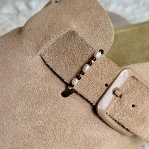 May include: A beige suede shoe strap adorned with a delicate beaded charm featuring alternating small white pearls, dark brown beads, and metallic gold spacers. The strap includes a matching beige buckle, showcasing a textured finish on the suede material.