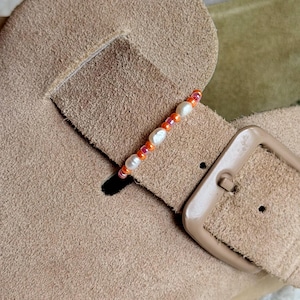 May include: A beaded accessory featuring a sequence of small, round, translucent orange beads interspersed with irregular, white, lustrous freshwater pearls, positioned across the suede strap of a tan-colored footwear item with a buckle.