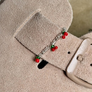 May include: A beige suede shoe strap adorned with a beaded charm featuring two pairs of small red and green cherry-shaped beads connected by a string of clear, translucent seed beads attached to the leather surface near a brown buckle.