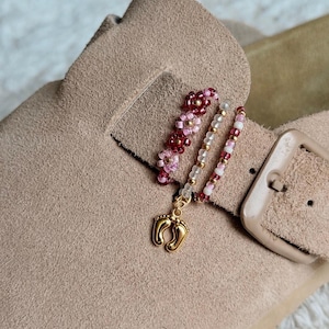 Könnte beinhalten: Ein beigefarbener Wildleder-Sandalenriemen, verziert mit zwei Perlenanhängern. Ein Anhänger besteht aus rosa und kastanienbraunen blumenförmigen Perlen, der andere aus einer Schnur aus rosa, weißen und goldenen Perlen mit einem goldenen Fußabdruck-Anhänger.