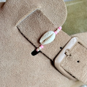 Könnte beinhalten: Ein beigefarbener Riemen aus Wildleder mit einem dekorativen Anhänger aus einer weißen Kaurimuschel, flankiert von kleinen rosa und weißen Perlen. Der Riemen verfügt über eine passende beigefarbene Schnalle und verläuft horizontal über den Schuh.