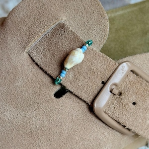 May include: A tan suede sandal strap featuring a decorative accent made of a small white seashell flanked by light blue and green beads. The strap includes a matching tan buckle and a rectangular cutout for the buckle prong to pass through.